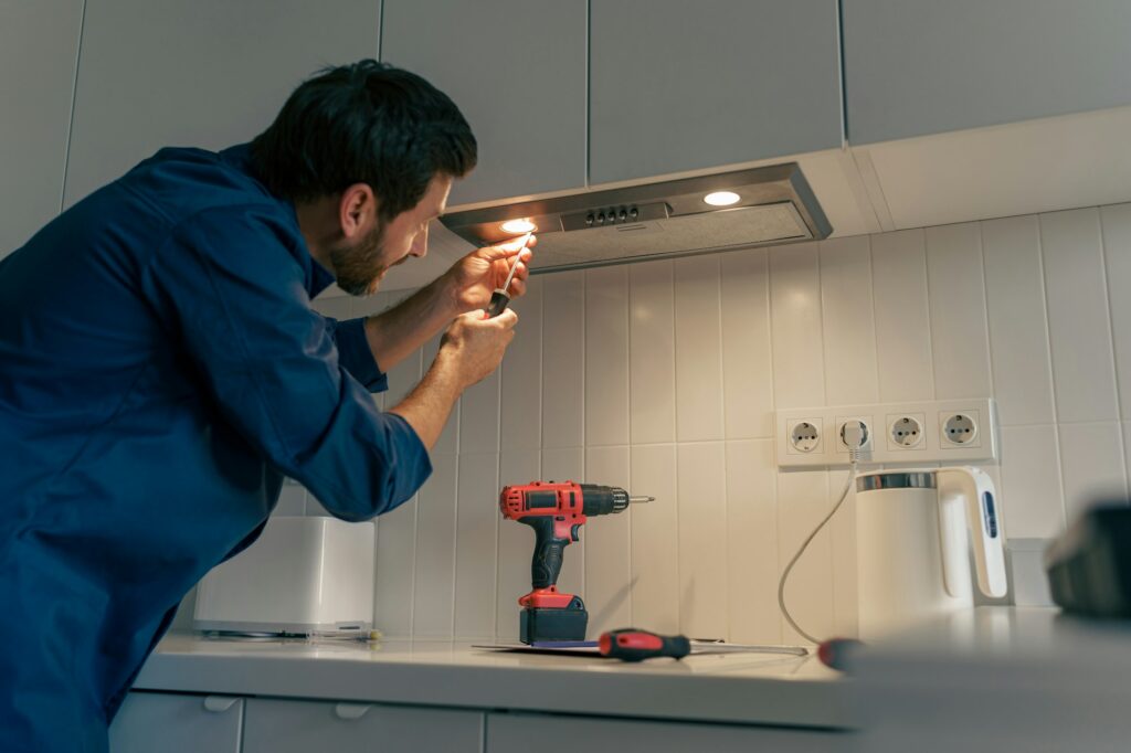Professional male worker in uniform repairing modern cooker hood in kitchen